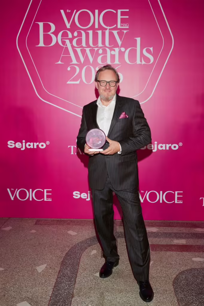мужчина в костюме и в очках, держит в руках награду VOICE Beauty Awards 2026